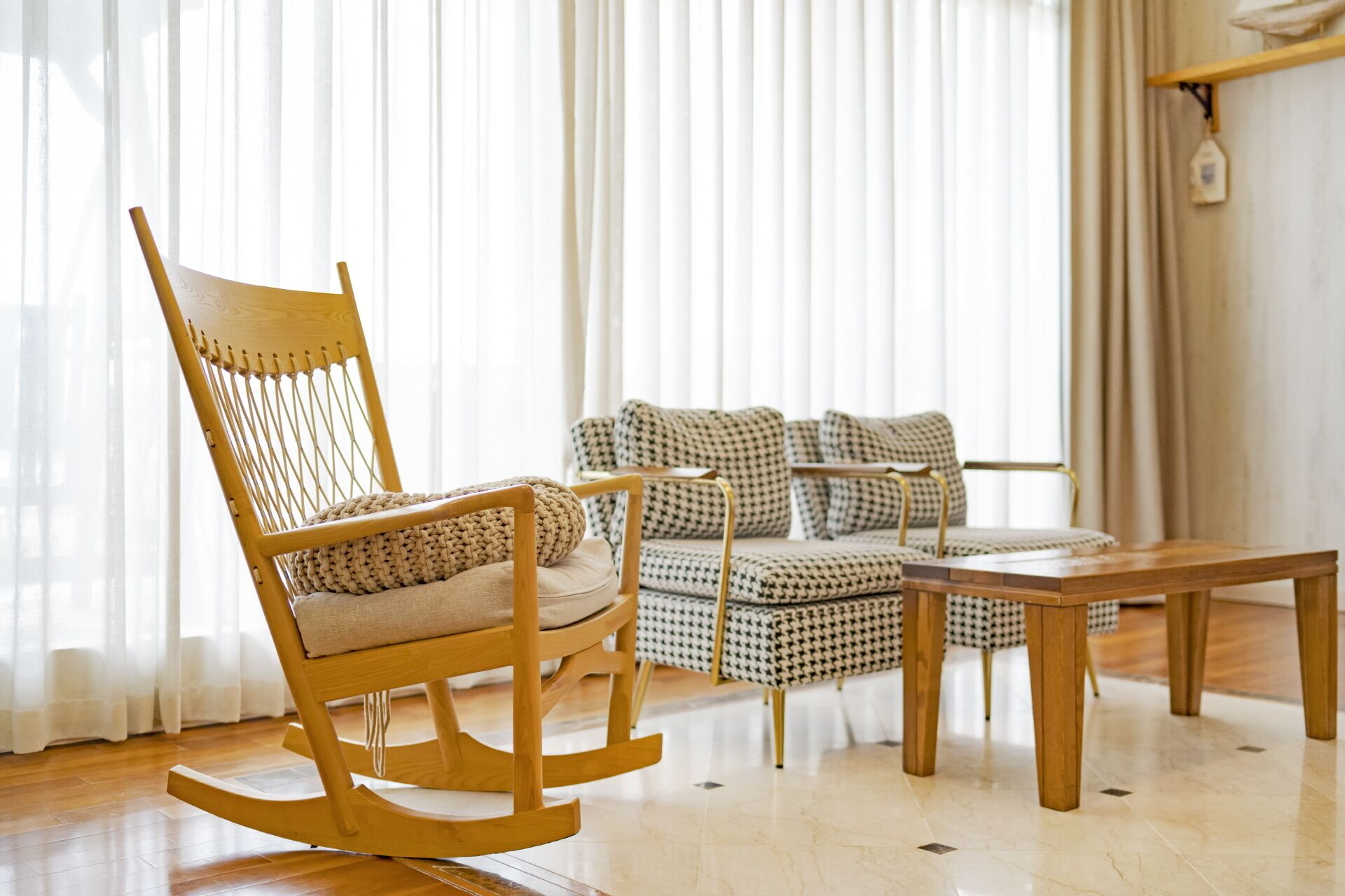 Un fauteuil à bascule en bois avec une assise et un dossier en osier, un canapé à motifs pied-de-poule et une table basse en bois dans une pièce dotée d’un plancher en bois et de rideaux légers.