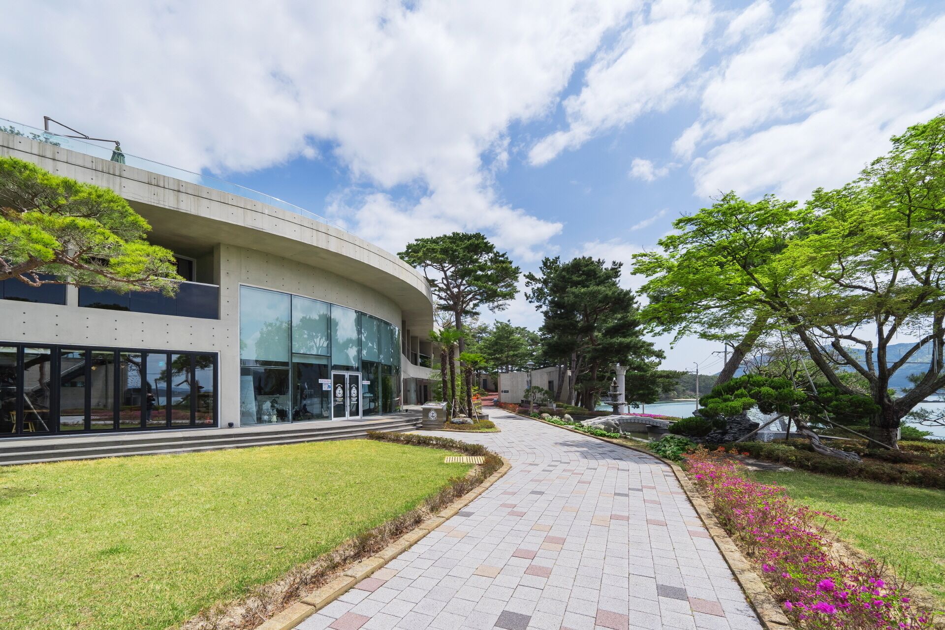 A modern building with a curved facade, a paved walkway, and a landscaped garden with trees and flowers.