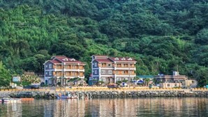 Un hotel frente al mar con edificios de varios pisos, un muelle con barcos y un paisaje de colinas cubiertas de bosque al fondo.