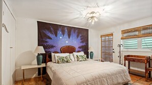 A bedroom with a large bed, two bedside tables, a nightstand, a ceiling light fixture, a window with blinds, and a wall tapestry. - Quiet and safe Uptown house steps from Audubon Zoo and shopping on Magazine (New Orleans)