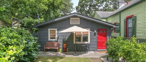 A small, single-story building with a red door, a bench, and an umbrella in front.