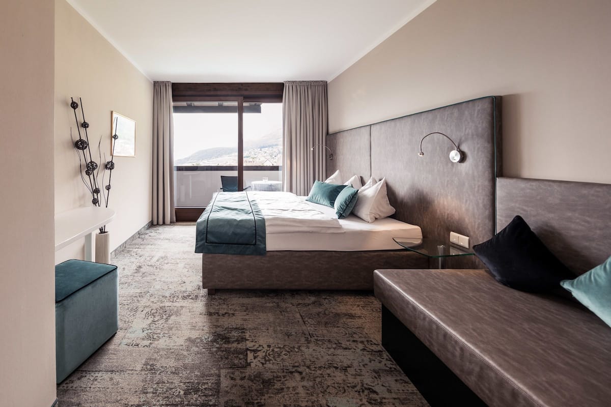 Ein modernes Hotelzimmer mit einem großen Bett, einer Couch, einem kleinen Tisch und Blick auf die Stadt durch große Fenster.