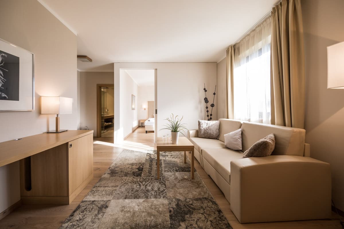 Ein modernes Wohnzimmer mit einer Couch, einem Couchtisch und einem Teppich. Es gibt einen Schreibtisch mit Lampe und Pflanze. Das Zimmer hat ein großes Fenster mit Vorhängen.