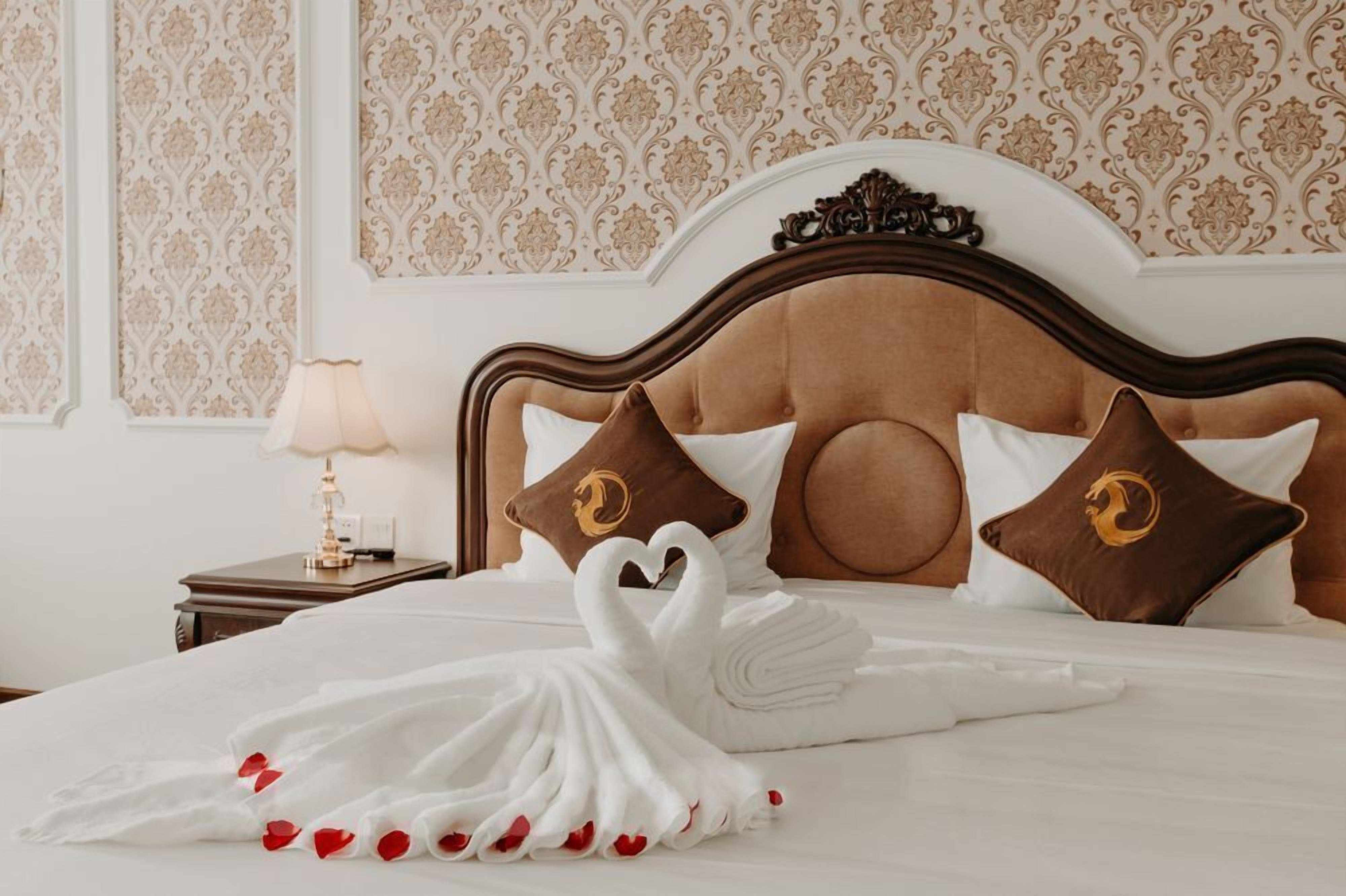 A neatly made bed with a swan towel arrangement and rose petals, a wooden headboard, and matching pillows with a crest emblem.