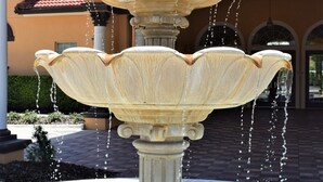 Une fontaine à plusieurs niveaux, dont l’eau jaillit en cascade sur chaque niveau, se dressant devant un bâtiment orné d’arcades et surmonté d’un toit en tuiles.