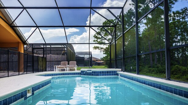 Une piscine intérieure à l’eau d’un bleu cristallin, entourée d’une structure de verre, avec vue sur une maison et des arbres en arrière-plan.