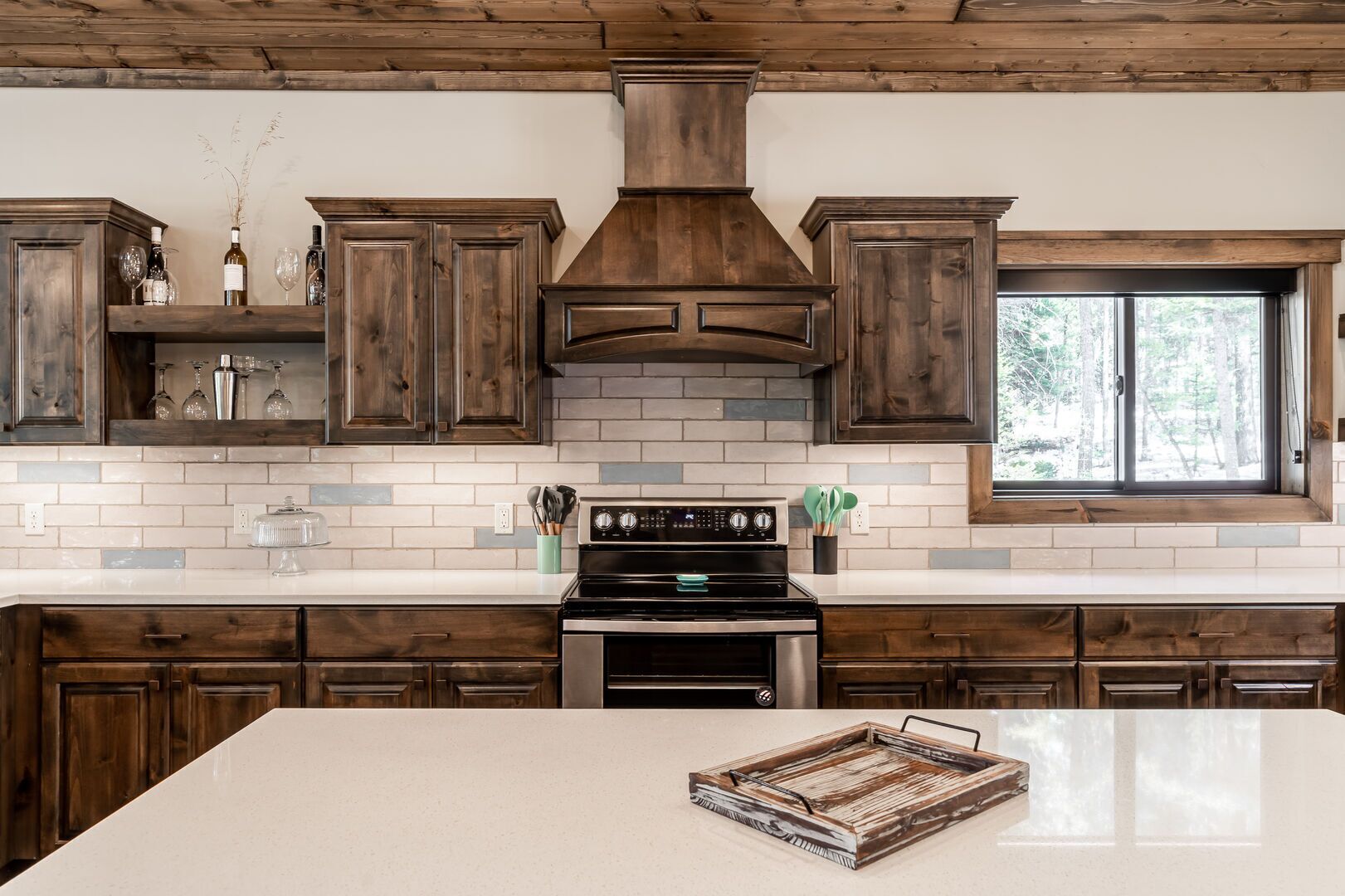 A kitchen with dark wooden cabinets, a white tiled backsplash, a stainless steel stove, and a window with a view of trees.