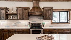 A kitchen with dark wooden cabinets, a white tiled backsplash, a stainless steel stove, and a window with a view of trees.