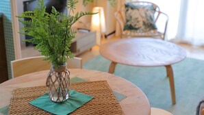 Une table ronde en bois, surmontée d’un set de table en fibres tressées, un vase en verre contenant des feuilles vertes et une chaise agrémentée d’un coussin à motifs.
