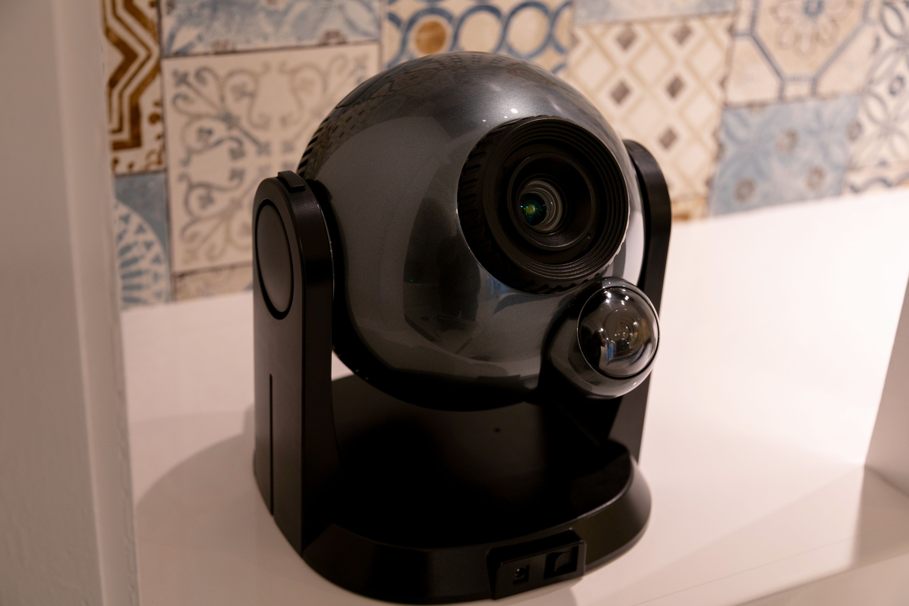 A surveillance camera with a dome design, positioned on a white surface against a tiled wall.