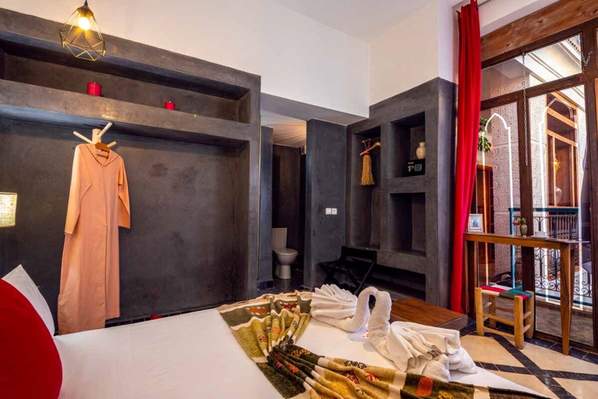 A hotel room with a bed, a hanging robe, a towel rack, a small table, and a view of the outside.