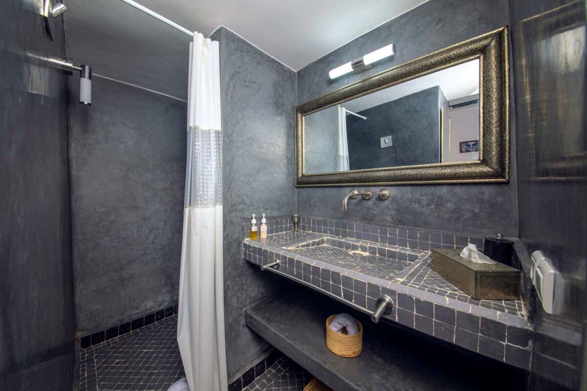 A modern bathroom with a dark tiled wall, a large mirror with a gold frame, a shower area, and a sink with a small trash bin.