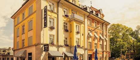Et hotel med flere etager, en gul facade, altaner med blomsterkasser og udendørs siddepladser.