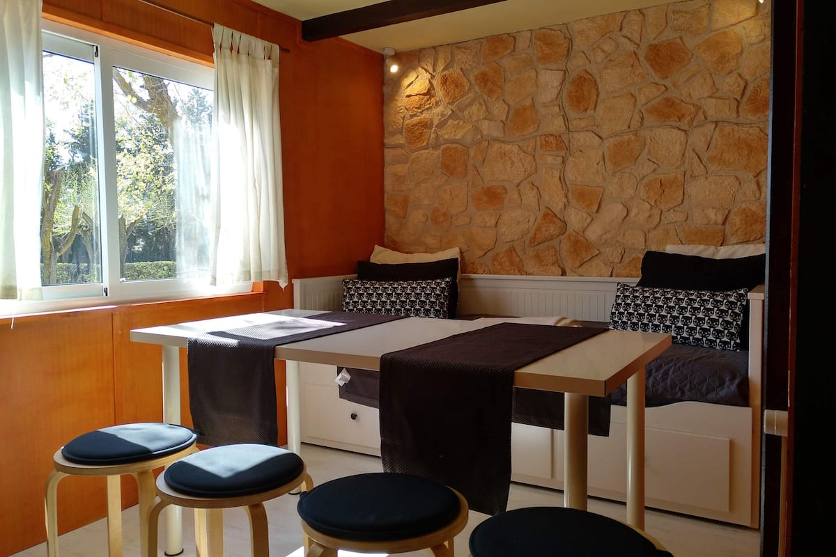 A cozy room with a stone wall, a bed with patterned pillows, a table with a tablecloth, and two stools.
