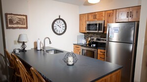 A kitchen with wooden cabinets, a stainless steel refrigerator, a microwave, and a clock on the wall. - Mountain Lodge At Okemo-1br Fireplace & Updated Kitchen 1 Bedroom Condo by RedAwning (Ludlow)