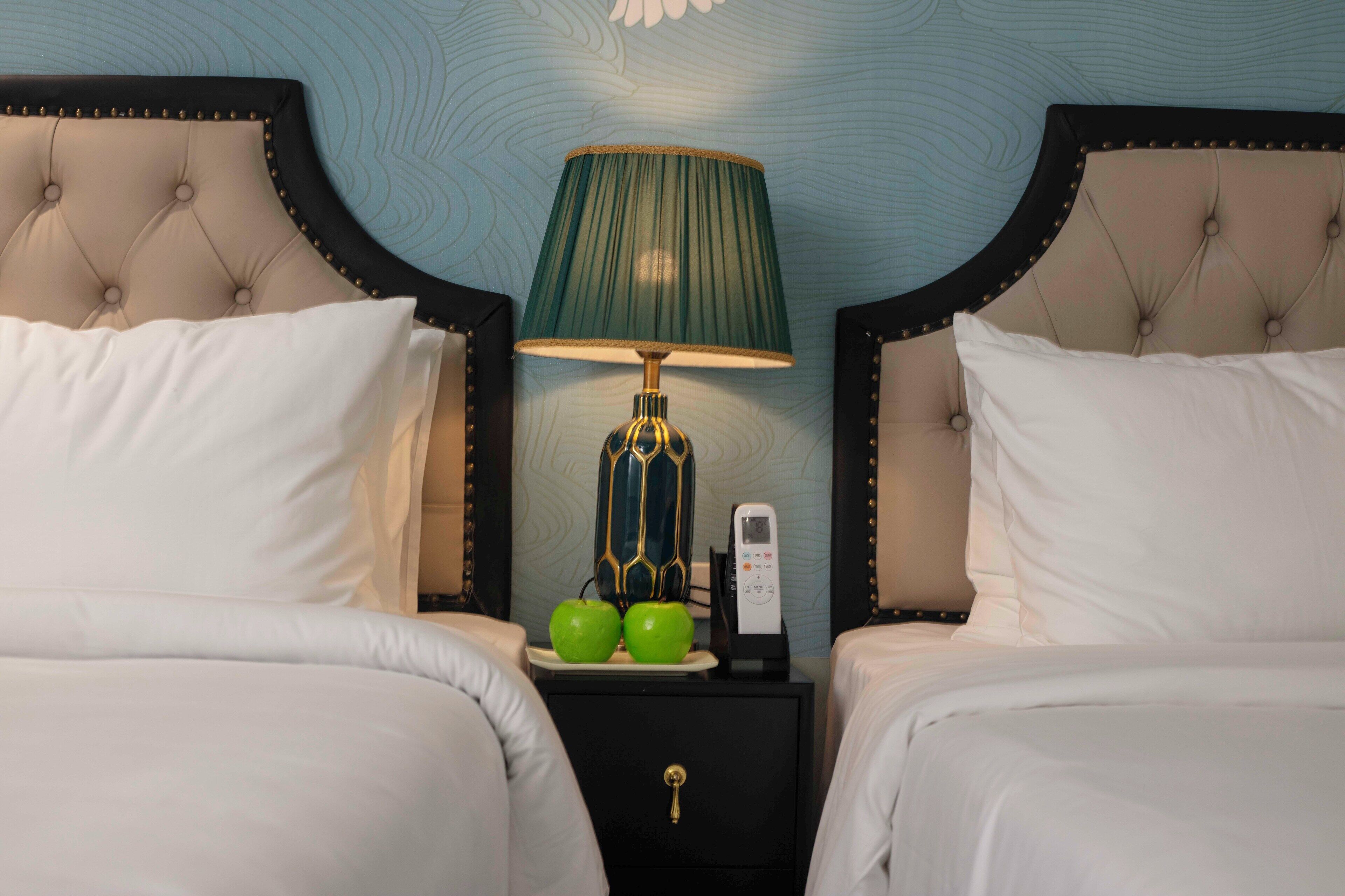 A hotel room with two beds, a nightstand with a lamp, two green apples, and a phone.