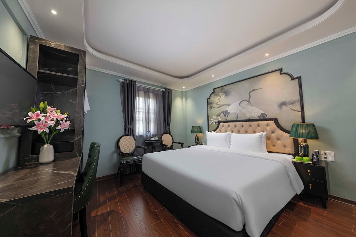 Une chambre d’hôtel avec un grand lit, une table de chevet avec une lampe, une chaise, une petite table avec un vase de fleurs et un tableau au mur.