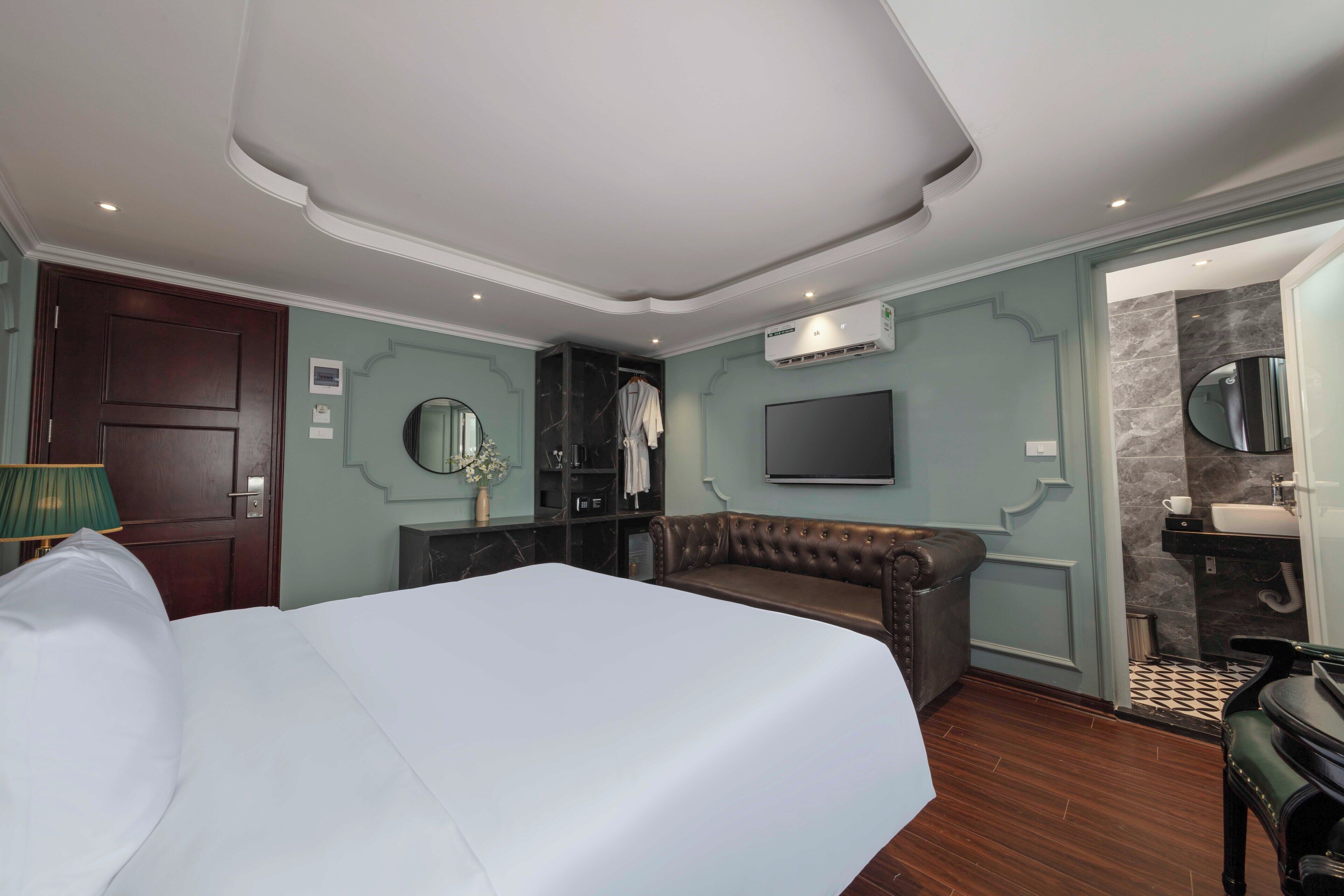 Habitación de hotel con una cama grande, un sofá de cuero, un televisor de pantalla plana, un baño con lavabo y espejo, y un panel decorativo en la pared.