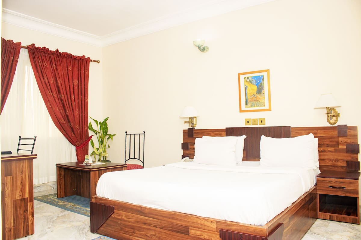 A hotel room with a wooden bed, a desk, a chair, a nightstand, and a wall-mounted lamp.