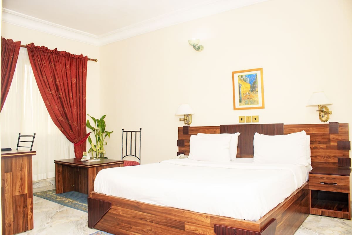 A hotel room with a wooden bed, a desk, a chair, a nightstand, and a wall-mounted lamp.