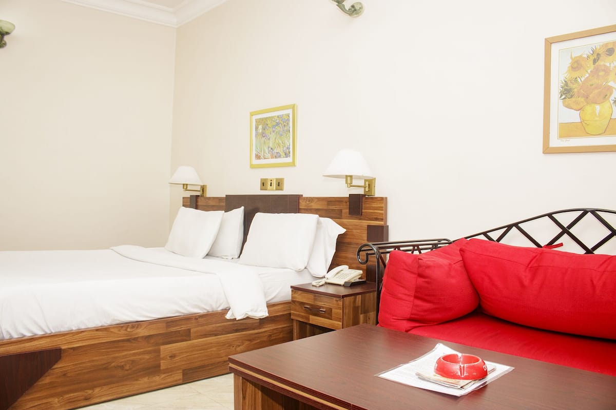 A hotel room with a wooden bed, a red sofa, a wooden nightstand with a lamp, and a framed artwork on the wall.