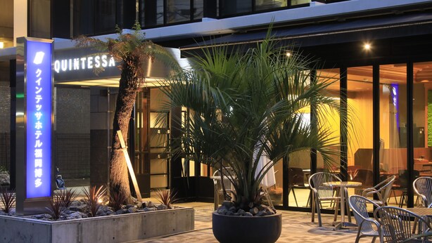 An outdoor seating area with chairs and a large potted plant, a sign for 'Quintessa', and a building entrance. - Quintessa Hotel Fukuoka Hakata Relax&Sleep (Fukuoka)