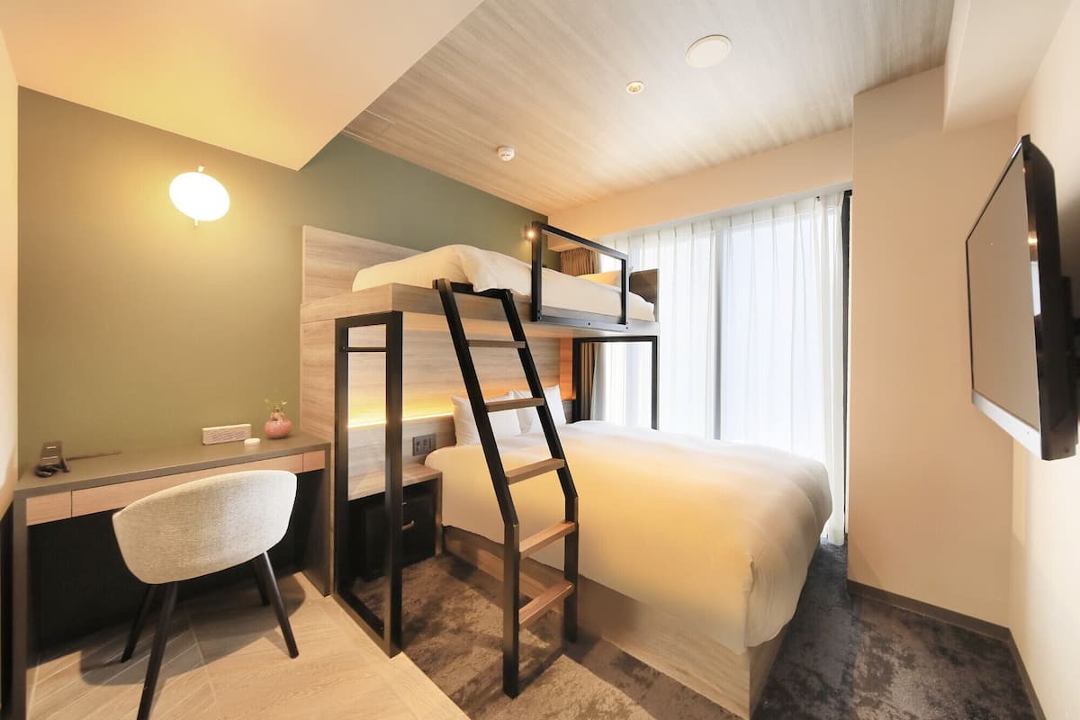 A hotel room with bunk beds, a desk, a chair, a TV, and a window with curtains.