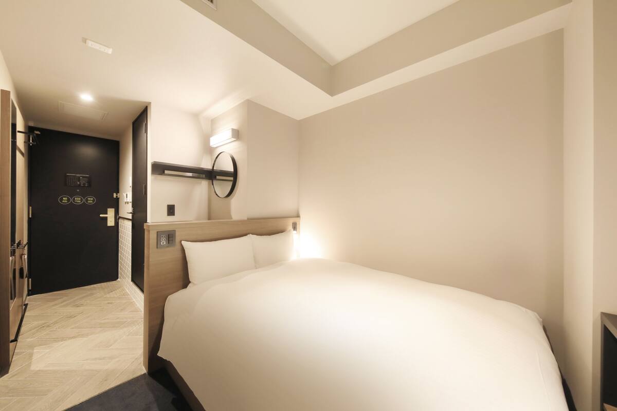 A modern hotel room with a large bed, a door with a control panel, and a wall-mounted light fixture.