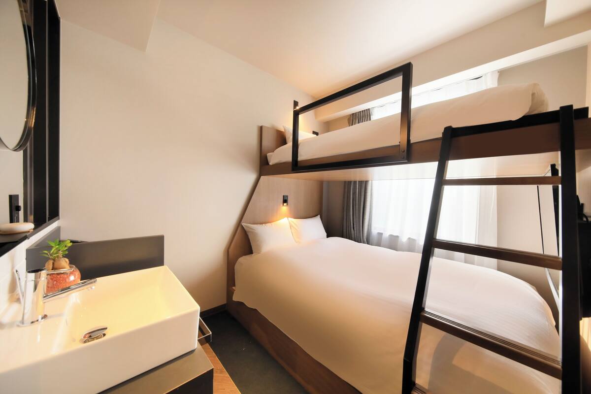 A hotel room with a bunk bed, a bedside table with a mirror, a small potted plant, and a window with curtains.