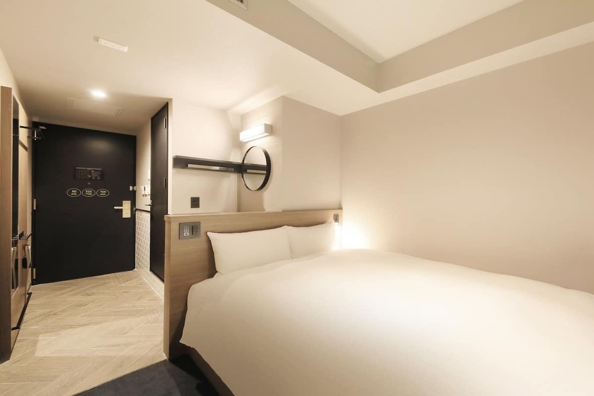 A hotel room with a large bed, a bedside table, a wall-mounted mirror, and a door with an electronic control panel.
