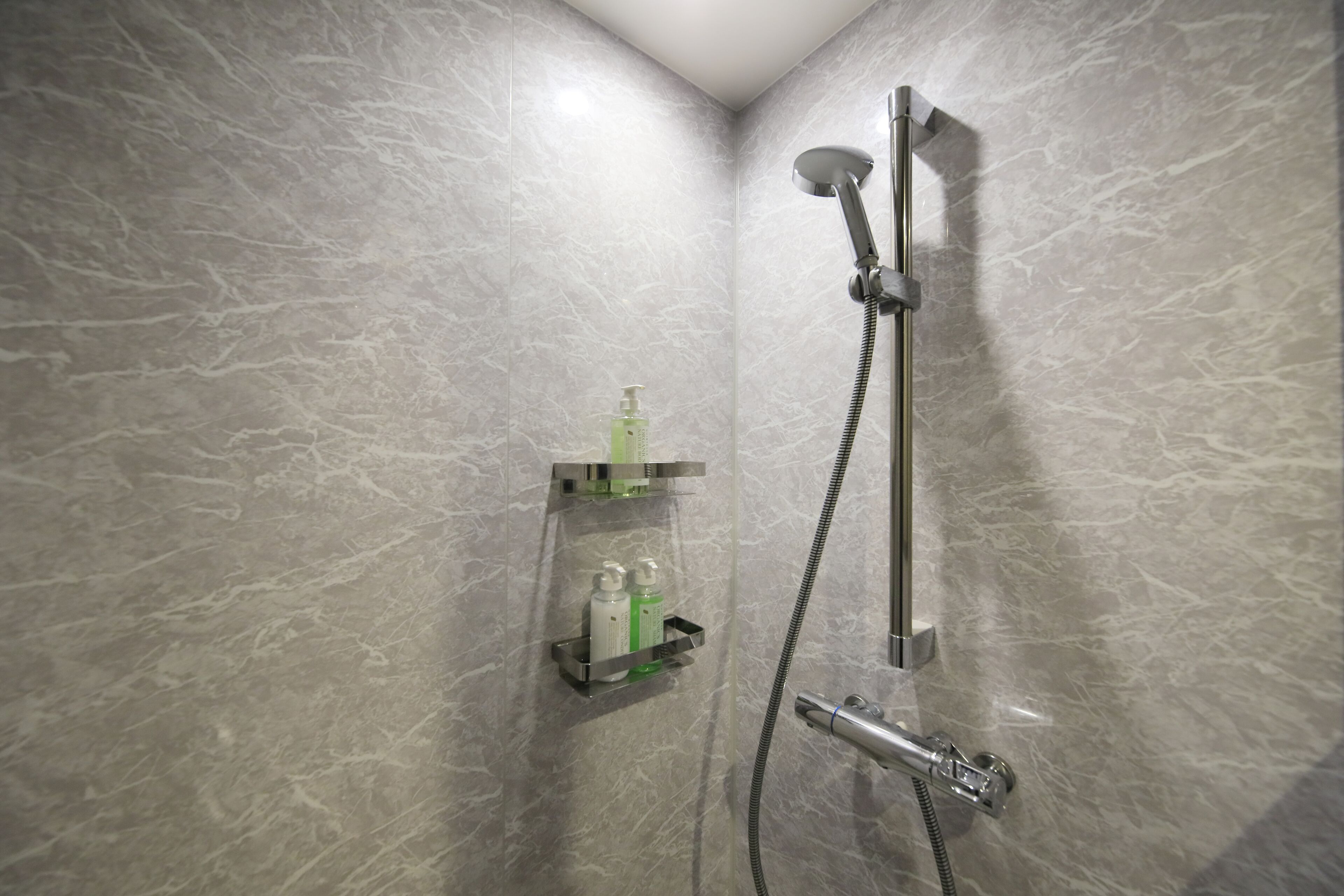 A modern shower with a handheld showerhead and a wall-mounted faucet. There is a shelf with toiletries.