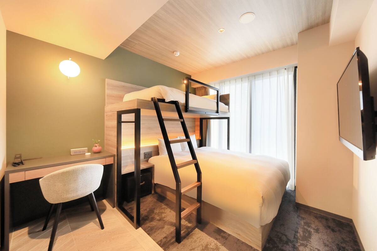 A hotel room with a bunk bed, a desk, a chair, a TV, and a window with curtains.