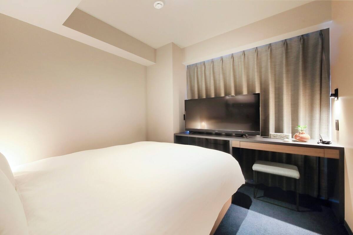 A hotel room with a bed, a desk with a TV, a chair, and a window with curtains.