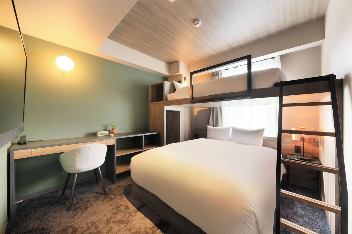 A hotel room with a bunk bed, a desk with a chair, a bedside table with a lamp, and a window with curtains.
