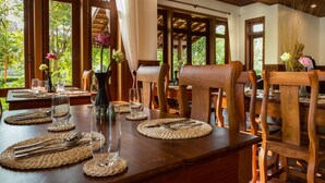 Un comedor con mesas y sillas de madera, dispuesto con cubiertos y cristalería, y ventanas amplias que ofrecen vista a la vegetación.