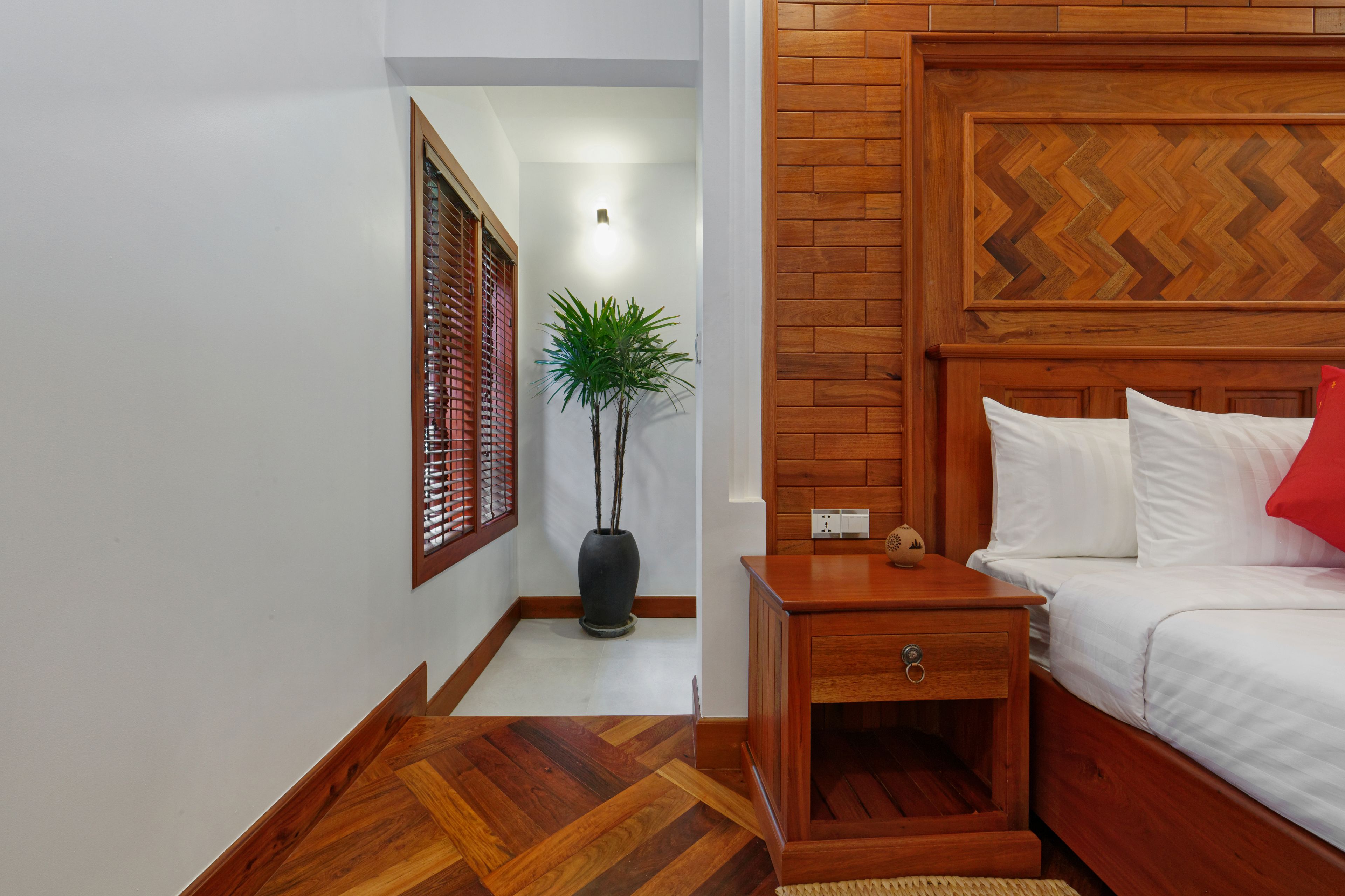 A bedroom with a wooden headboard, a bedside table, a nightstand, a potted plant, and a bed with white bedding and a red pillow.