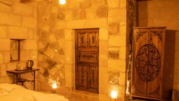A stone-walled room with a wooden door, a cabinet with intricate carvings, a small table with a kettle, and a bed with white linens.