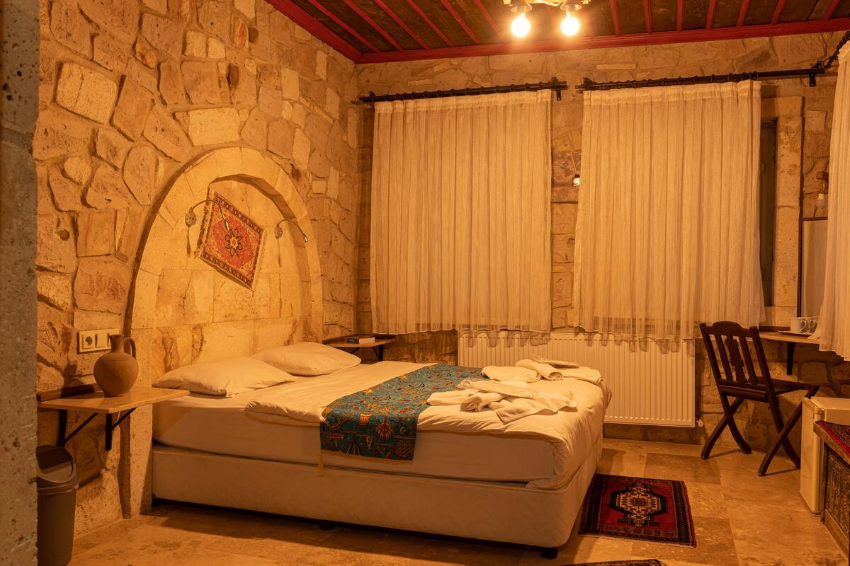 A bedroom with a stone wall, a bed with a patterned blanket, a wooden chair, and a small table with a cup.