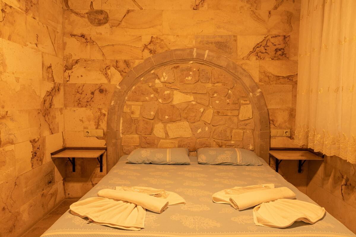 A room with a stone wall, a bed with a headboard, two bedside tables, and towels on the bed.