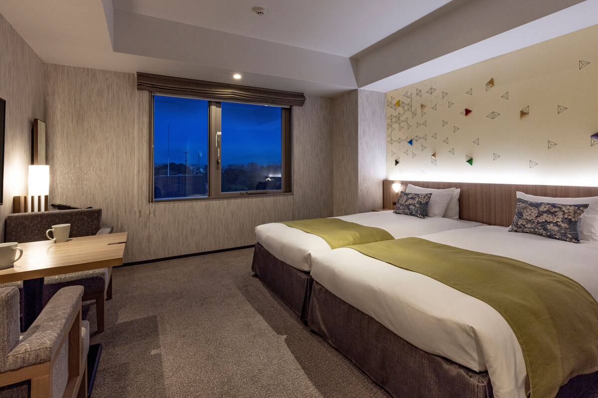 A hotel room with a large bed, a desk with a chair, a window with a view, and a wall with geometric patterns.