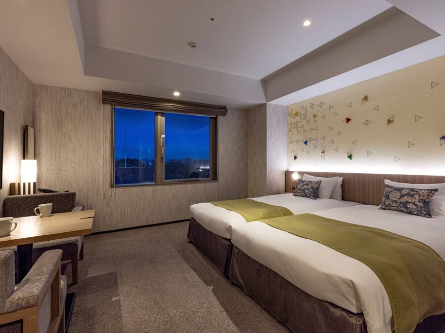 A hotel room with a large bed, a desk with a chair, a window with a view, and a wall with geometric patterns.
