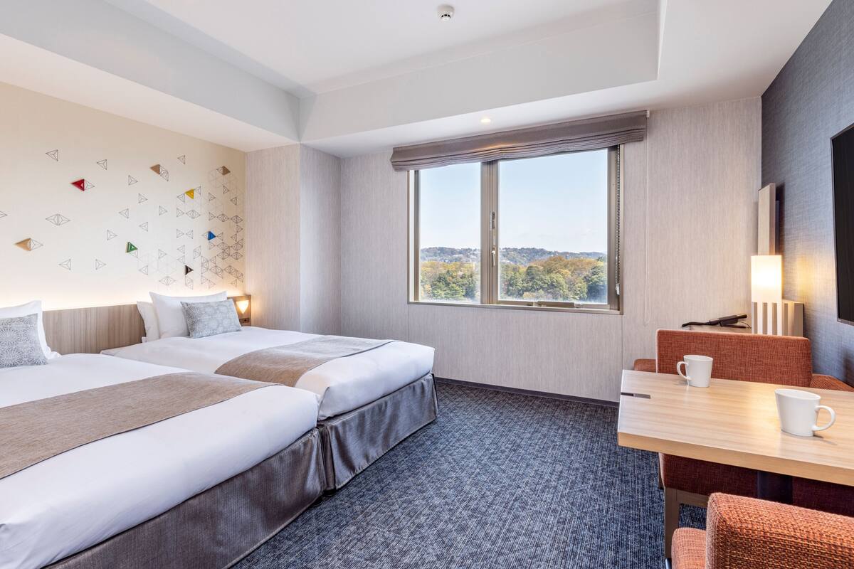 A hotel room with two beds, a desk, a chair, a window with a view, and a wall with geometric patterns.