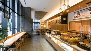 Interior de cafè modern amb taules i cadires de fusta, un mostrador de buffet amb diferents plats, i un gran finestral que ofereix vista al carrer.