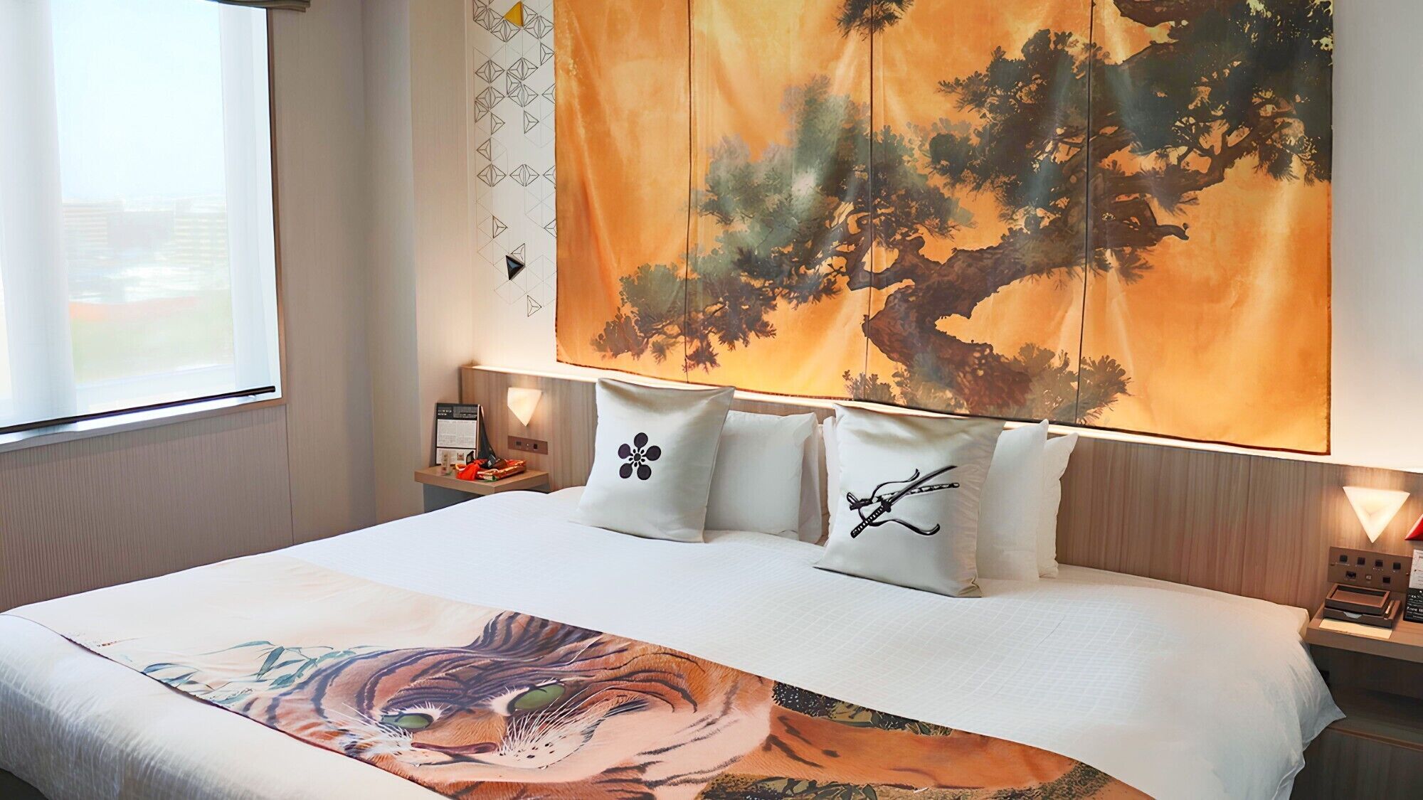 A hotel room with a large bed, a window with a view, a nightstand with a lamp, and a wall with a tree painting.