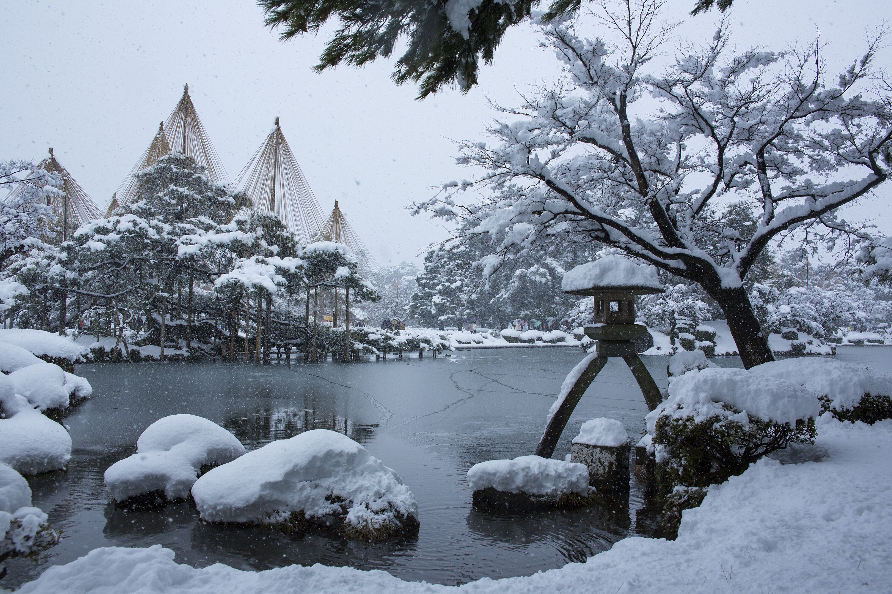 Escena invernal serena con un paisaje cubierto de nieve, que incluye un estanque, árboles cargados de nieve y construcciones tradicionales al fondo.