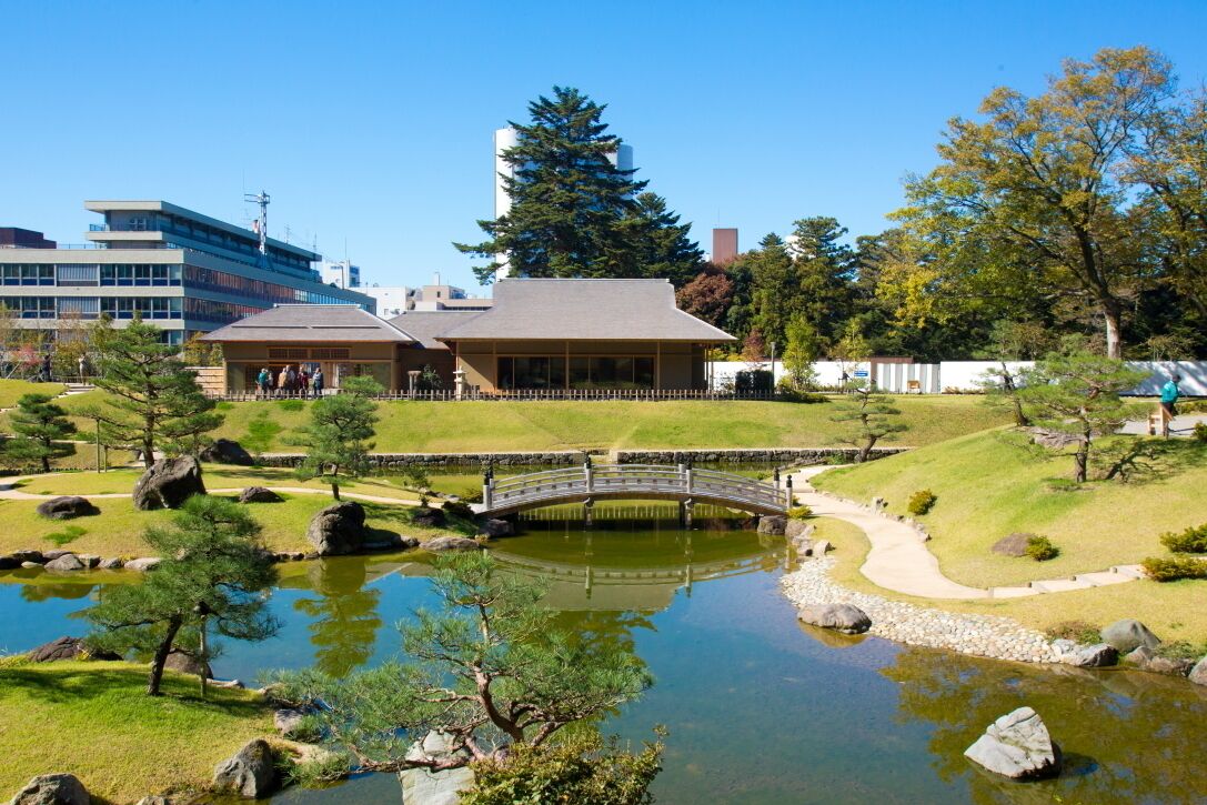 A traditional Japanese garden with a pond, a stone bridge, and a building with a sloped roof.