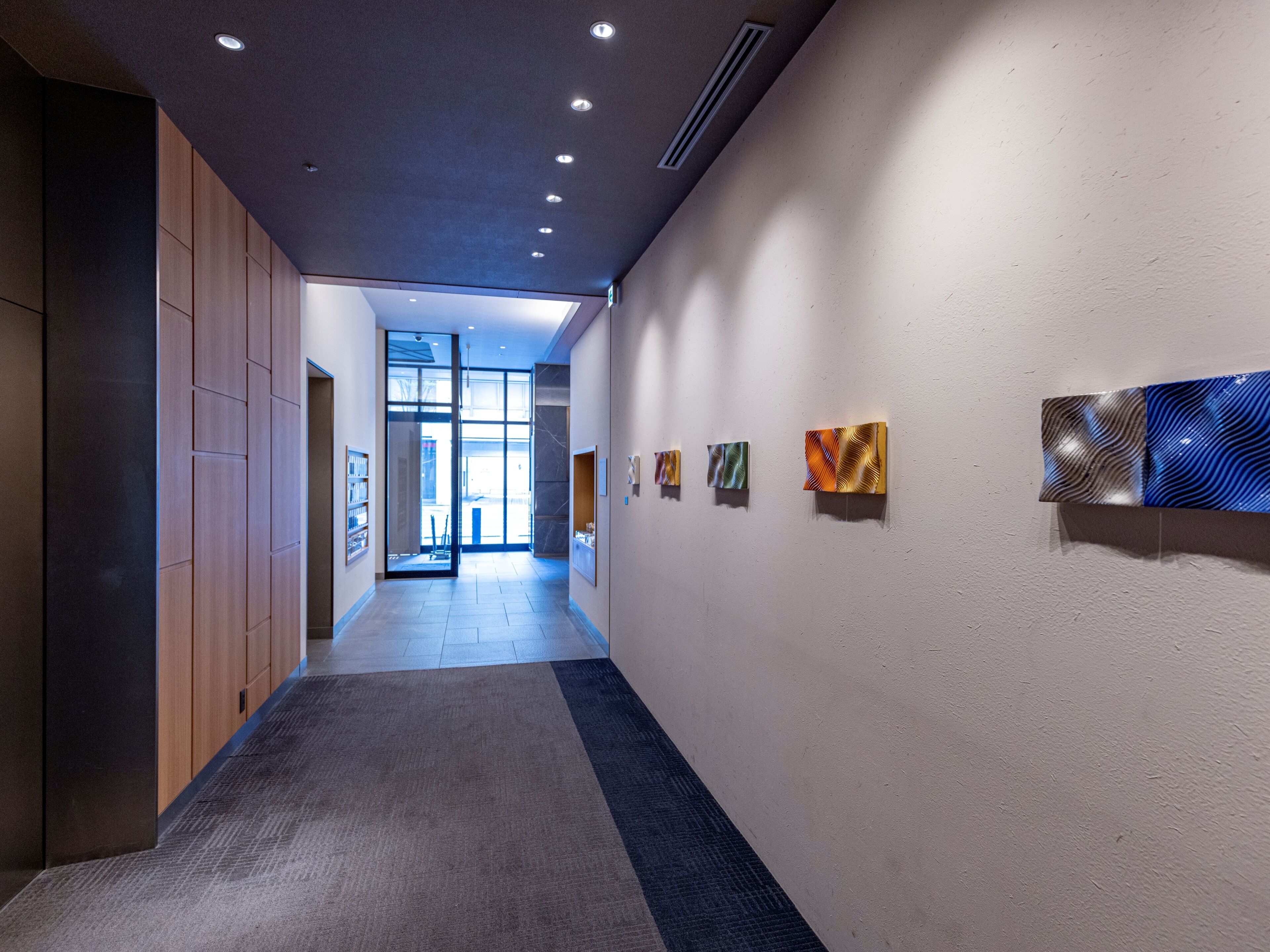 A hallway with a carpeted floor, wooden paneling on the left, and a series of artworks on the right wall.