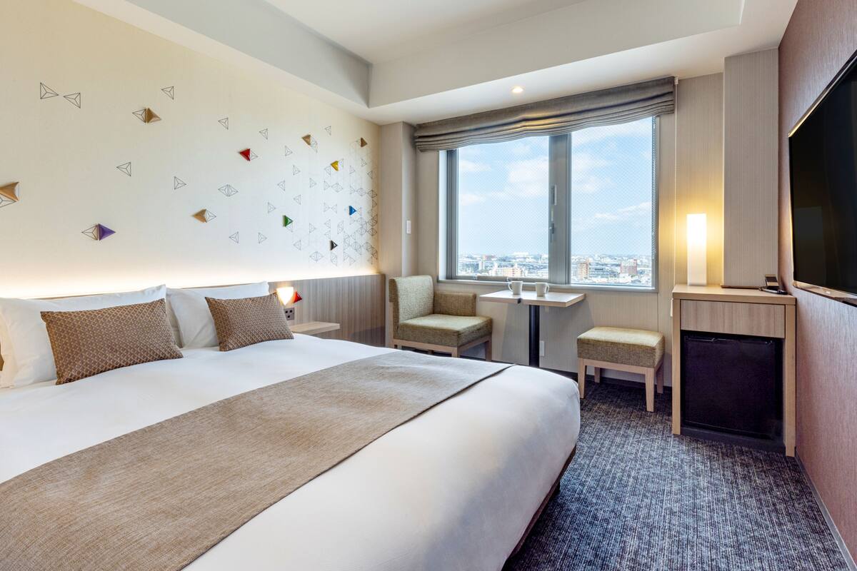 A hotel room with a large bed, a small table, two chairs, a TV, and a window with a city view.