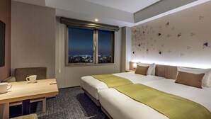 Habitació d'hotel amb un llit gran, una taula amb dues cadires, una finestra que dona a una vista de la ciutat i una paret amb dibuixos geomètrics.