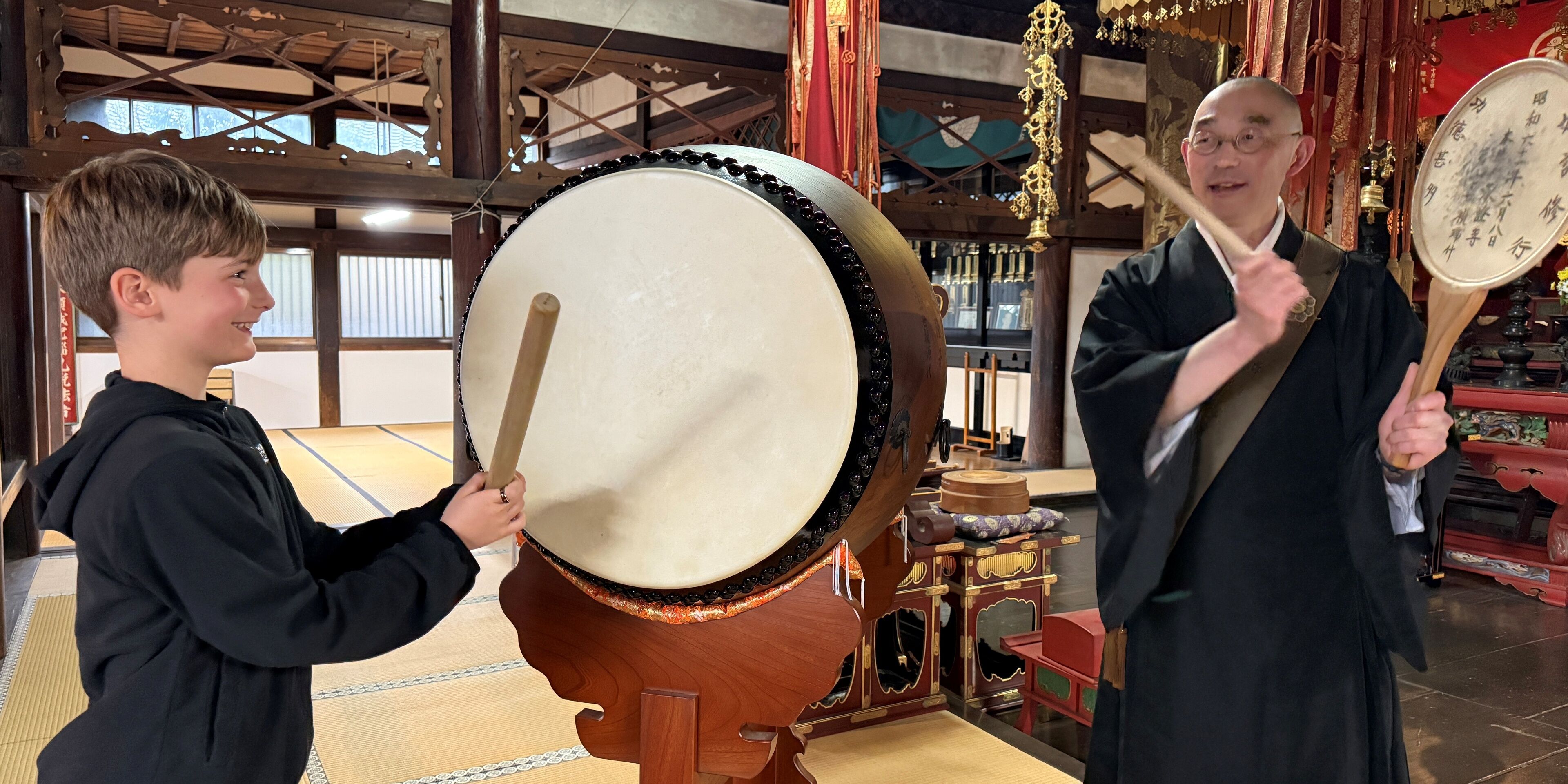 Two individuals playing large drums in a room with traditional decor and wooden furniture.